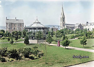 An old image that has been restored and colourised of Warrenpoint park and its bandstand with St Peter's RC church in the background